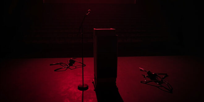 Dark university auditorium with single microphone on podium, empty chairs scattered around, dramatic shadows
