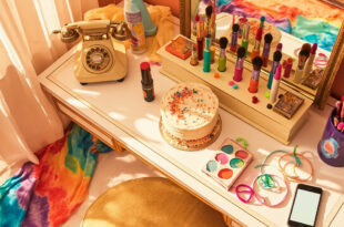 Vintage rotary phone beside colorful makeup palette, tie-dye clothing, and golden cake on vanity table