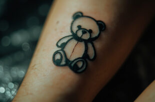 Delicate black ink teddy bear tattoo on forearm in professional tattoo studio setting