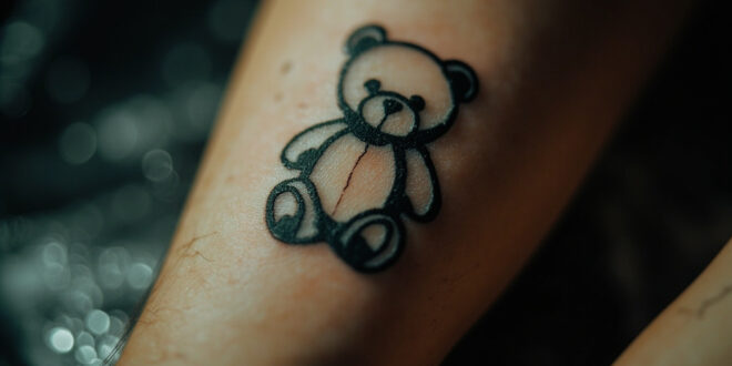 Delicate black ink teddy bear tattoo on forearm in professional tattoo studio setting