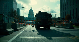 Military checkpoint with armored vehicle at state boundary, warning barriers, and constitutional documents near government buildings.