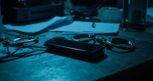Smartphone and handcuffs on police evidence table with case files and forensic equipment in background