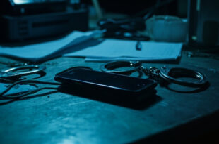 Smartphone and handcuffs on police evidence table with case files and forensic equipment in background