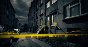 Yellow police tape blocks entrance to gray apartment building with emergency vehicles visible in foreground