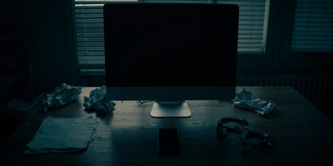 Dark home office with closed laptop, scattered papers, and handcuffs on wooden desk