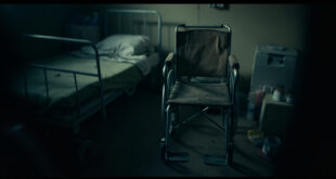 Empty wheelchair beside hospital bed in small apartment room with medical supplies and ventilator nearby