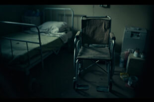 Empty wheelchair beside hospital bed in small apartment room with medical supplies and ventilator nearby