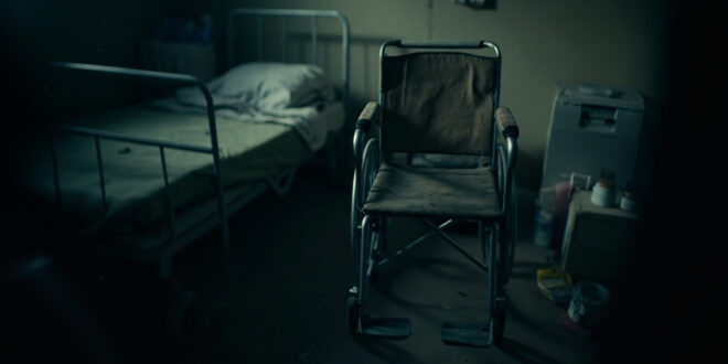 Empty wheelchair beside hospital bed in small apartment room with medical supplies and ventilator nearby