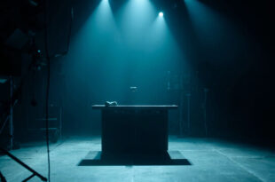 Empty television studio desk with microphone under spotlight, darkened screens and broadcasting equipment in background
