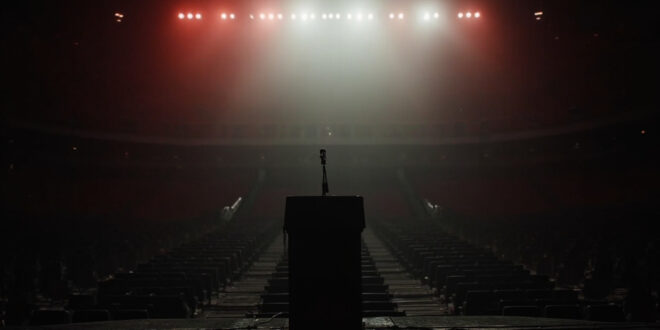 Empty stadium stage with lone microphone under bright spotlights, rows of vacant seats extending into darkness