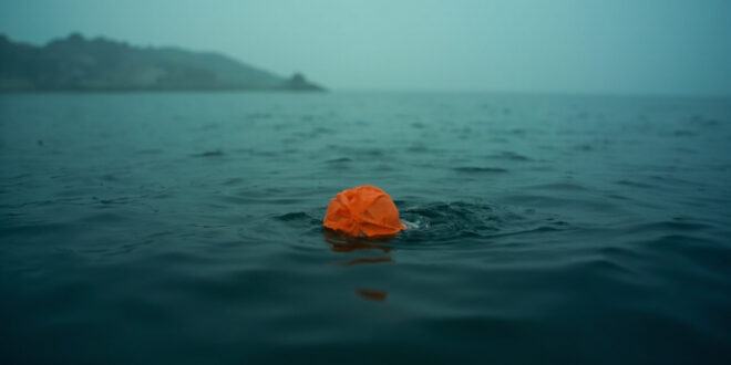 Orange swim cap floating alone on dark bay waters with misty shoreline in background