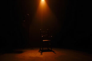 Empty black chair at podcast recording desk under focused studio lighting in darkened room