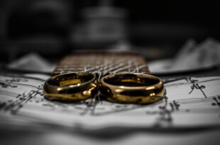 Two golden wedding rings rest on acoustic guitar strings in a modest apartment setting with medical recovery items nearby