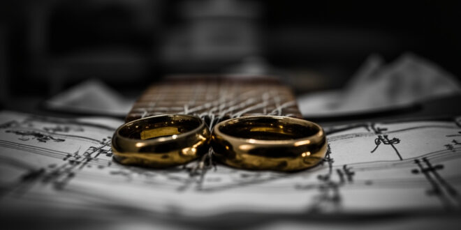Two golden wedding rings rest on acoustic guitar strings in a modest apartment setting with medical recovery items nearby