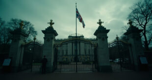 Ornate palace gates with the Union Jack flag at half mast against an overcast sky, with an empty announcement board