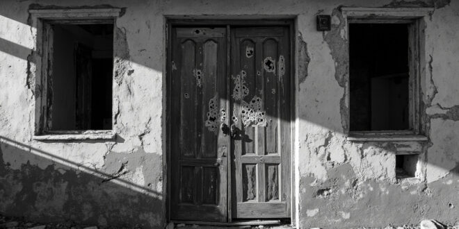 Weathered wooden door with bullet damage in abandoned desert building, harsh sunlight casting long shadows