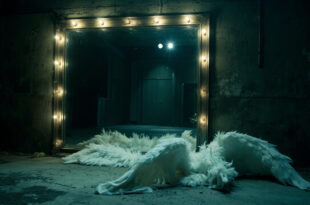White feathered angel wings leaning against gray concrete wall in empty backstage corridor with dramatic shadows