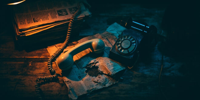 Vintage telephone handset rests beside crumpled newspapers on weathered wooden surface with dramatic shadows