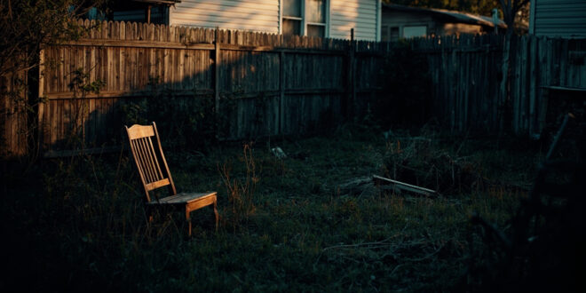 Empty suburban backyard with overturned furniture and overgrown grass beside weathered fence in fading light