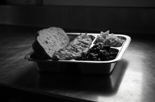 Institutional meal tray with baked fish, vegetables, and bread on stark surface under fluorescent light