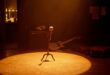 Empty wooden stage with vintage microphone stand under warm spotlight, guitar case and amplifier visible in shadows