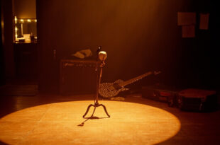 Empty wooden stage with vintage microphone stand under warm spotlight, guitar case and amplifier visible in shadows