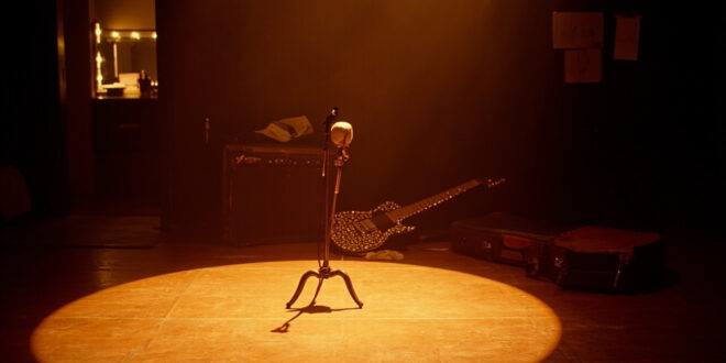 Empty wooden stage with vintage microphone stand under warm spotlight, guitar case and amplifier visible in shadows
