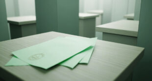Green ballot papers arranged on wooden surface next to transparent ballot box in polling station