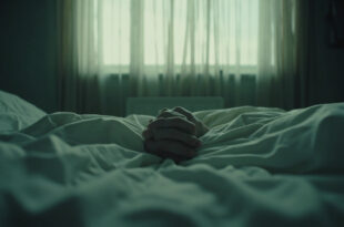 Two hands clasped together on white hospital bedding in a quiet medical room.