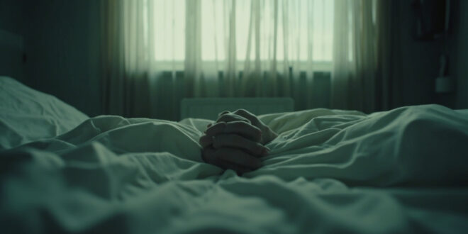 Two hands clasped together on white hospital bedding in a quiet medical room.