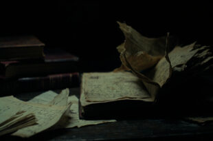 Open book with torn pages scattered on dark wooden desk beside ink-stained quill and crumpled letters