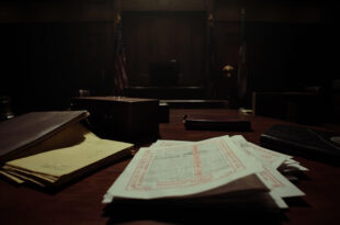 Closed briefcase with stacked legal documents and yellow notepad on dark wooden courtroom table in dramatic lighting
