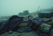 Half-moon glasses and flowing robes draped over driftwood on a foggy beach with film equipment nearby
