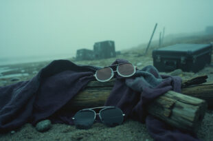 Half-moon glasses and flowing robes draped over driftwood on a foggy beach with film equipment nearby