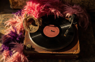 Pink feathered headpiece sits atop vintage record albums surrounded by theatrical costume pieces in golden lighting