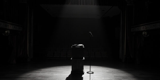 Empty speaker podium with open book under spotlight in classical opera house with burgundy curtains and vacant seats