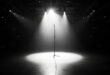 Single microphone stand illuminated by spotlight on empty stadium stage with rows of vacant seats in darkness