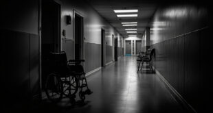 Empty wheelchair beside doorway in dimly lit hospital corridor with closed doors and fluorescent lighting overhead