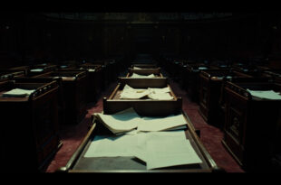 Scattered official documents and folders on dark wooden parliamentary desk in empty government chamber