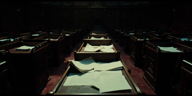 Scattered official documents and folders on dark wooden parliamentary desk in empty government chamber