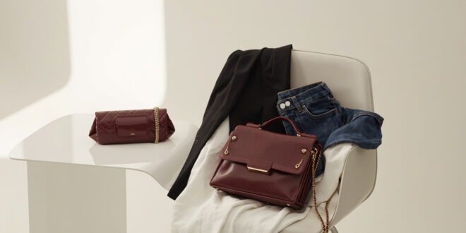 Burgundy leather handbag with gold chain beside white fabric and blazer on neutral minimalist background