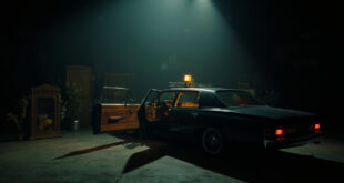 Open car door on dark soundstage with dramatic lighting and scattered memorial flowers nearby