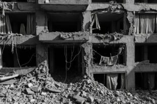 Concrete apartment building with shrapnel damage, broken windows, and large hole in upper floor surrounded by rubble