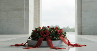 Ceremonial wreath with ribbon stands before monumental stone columns of a state memorial under gray sky
