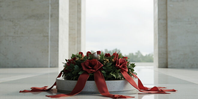 Ceremonial wreath with ribbon stands before monumental stone columns of a state memorial under gray sky