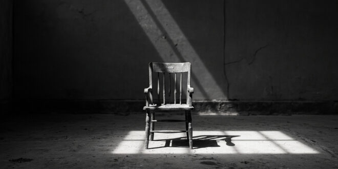 Single wooden chair alone in empty gray room with harsh shadows on bare concrete floor