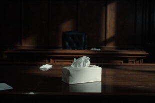 White tissue box sits on polished wooden table in empty courtroom with legal papers nearby