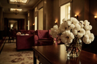 White cotton plant stems in clear glass vase on dark wooden table in hotel hallway with warm lighting