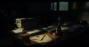 Official documents with stamps and seals lie on a wooden desk beside a broadcast microphone in dim office lighting