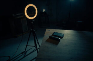 Dark recording studio with inactive ring light, smartphone on desk, microphone stand casting long shadow in empty room